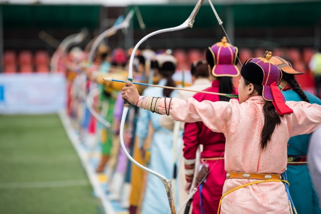 Women in traditional Mongolian dress lined up shooting bows and arrows.
