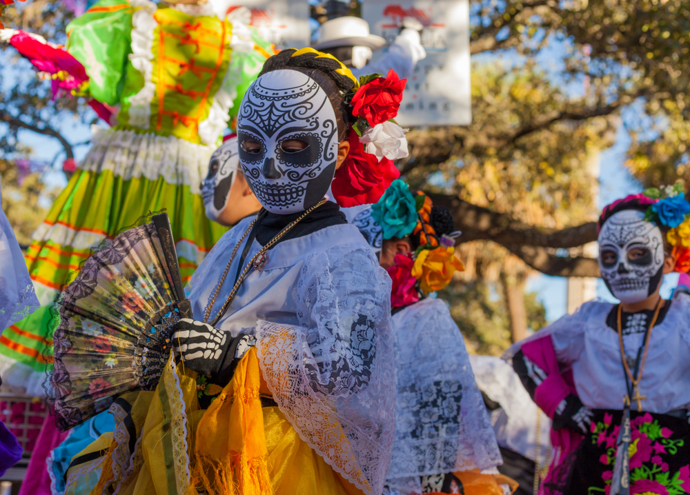 People dressed in colorful traditional outfits and skull face paint celebrate Day of the Dead, holding fans and flowers during a lively outdoor parade.