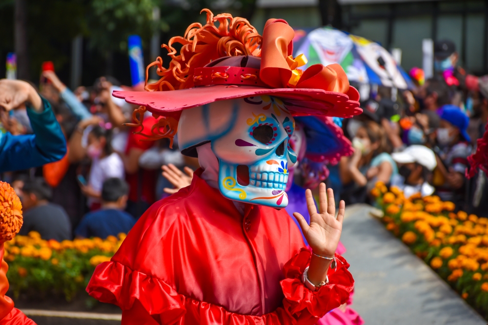 Person wearing colourful Day of the Dead costume with skull mask and red hat during a festive parade, surrounded by people and marigold flowers.