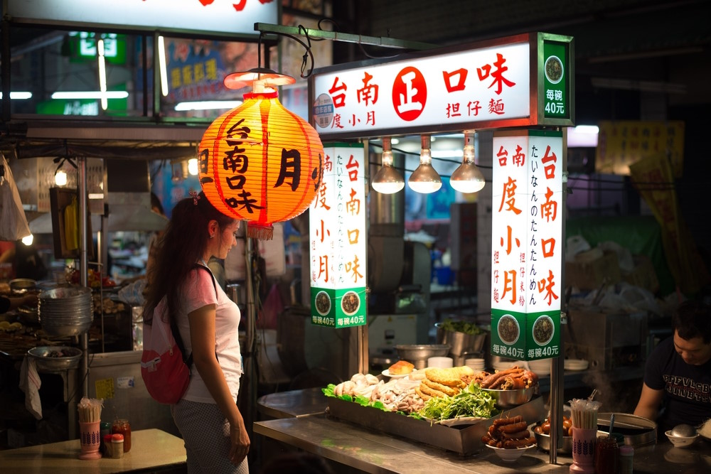 Taiwan night market food stall lit by red lanterns, Chinese signs overhead, trays of braised meats and greens, woman with backpack browsing at dusk.
