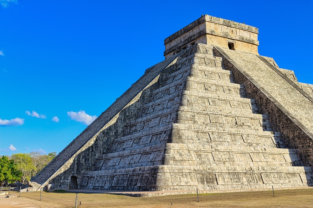 Ancient stone pyramid of Chichen Itza rises above a grassy plaza under a bright blue sky, showcasing the grandeur of Maya civilization.