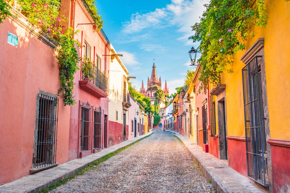 Cobbled street lined with colourful colonial buildings leads toward the iconic cathedral of San Miguel de Allende beneath a bright Mexican sky.