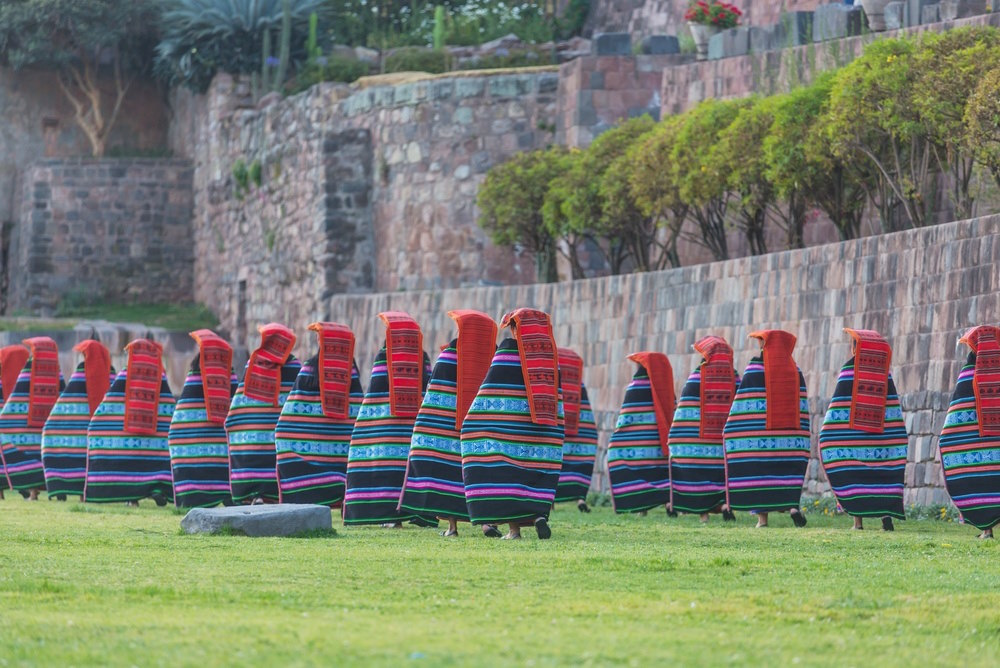 Performers in traditional Incan costume parade in front of the grey stone walls of an old Incan fortress for Inti Raymi festival